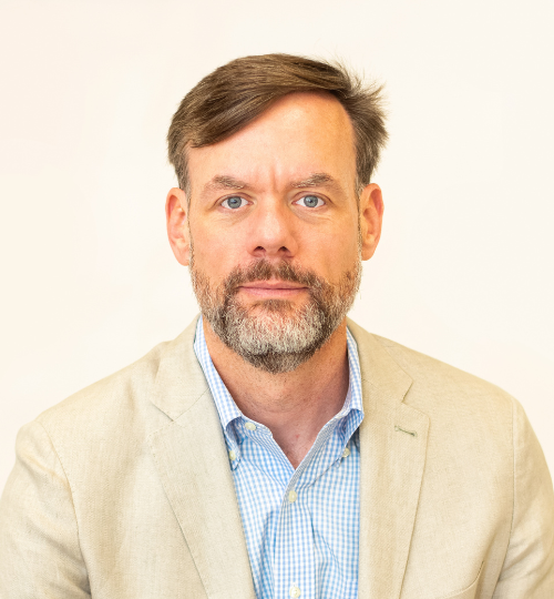 A middle aged man with brown hair and a greying goatee, wearing a blue and white striped Oxford button down shirt and a tan blazer over top, smiling for the camera.